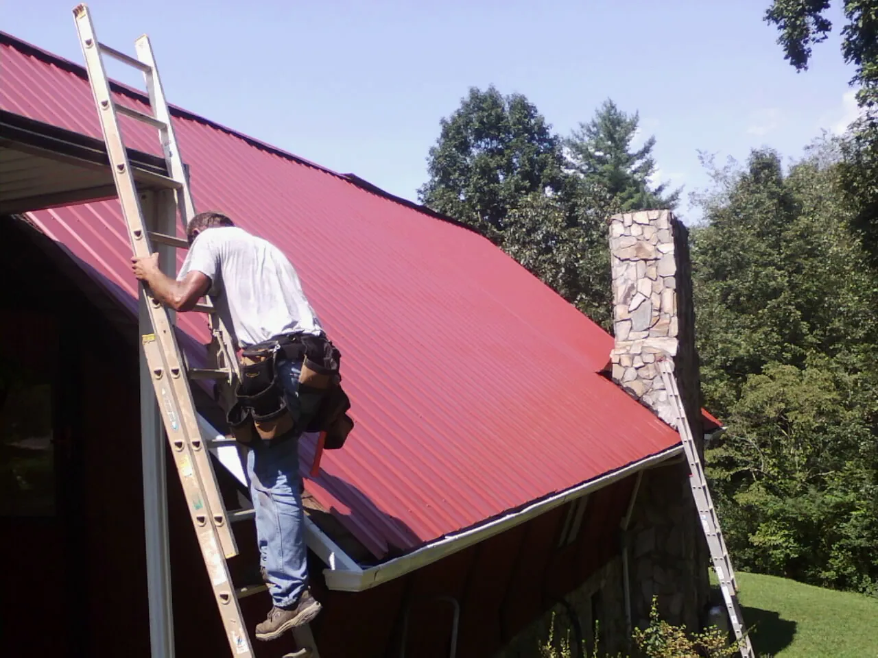 Roofer on a ladder beside a red metal roof and stone chimney — Soffit & Fascia Repair by Roofing Brothers in Brownville